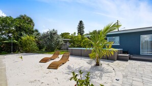 Un espacio exterior moderno con un patio arenoso, sillones de madera y una palmera.