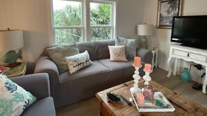 A living room with a grey sofa, a coffee table with candles, a television, and a window with a view of greenery. - 200ft of Waterfront with Tiki & Heated Pool - Boaters Paradise  (Key Largo)