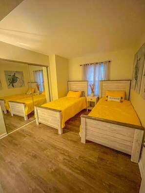 Une chambre avec trois lits simples, chacun recouvert d’une couvre-lit jaune et agrémenté d’oreillers assortis. Un grand miroir orne l’un des murs, une fenêtre est munie de rideaux, et un tableau représentant une tortue est accroché au mur opposé.