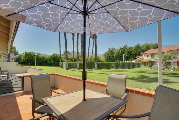 Une terrasse avec une table et des chaises, un parasol, et une vue sur une pelouse bien entretenue et des bâtiments situés au loin.