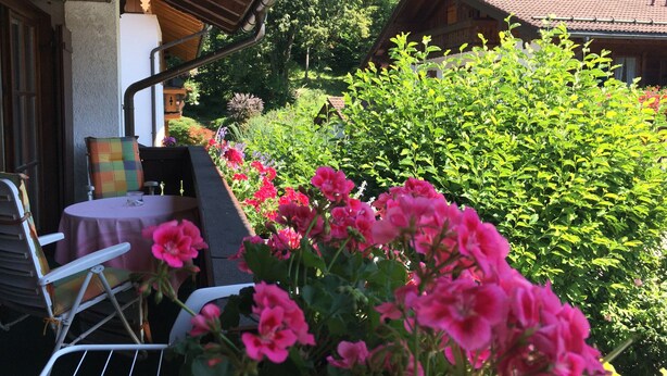 A patio with a table and chairs, surrounded by lush greenery and flowering plants. - Vacation apartment for 2 - 5 persons, approx. 85 m², living/sleeping room, 2 bedrooms, kitchenette, 2 bathrooms, 2 balconies, terrace (Bischofswiesen)