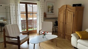 A living room with a wooden chair, a white sofa, a small table with a vase, a wooden cabinet, and a view through a glass door to the outside. - Vacation apartment for 2 - 5 persons, approx. 85 m², living/sleeping room, 2 bedrooms, kitchenette, 2 bathrooms, 2 balconies, terrace (Bischofswiesen)