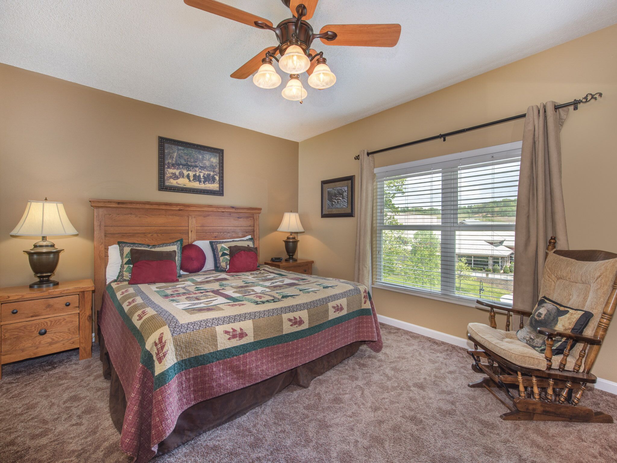 A bedroom with a wooden bed, two bedside tables with lamps, a ceiling fan, a rocking chair, and a large window with blinds.