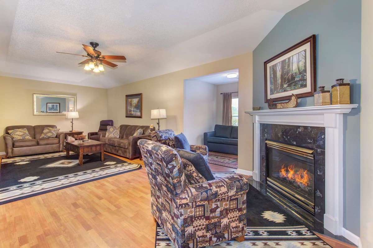 A living room with a fireplace, two sofas, a patterned armchair, and a ceiling fan.