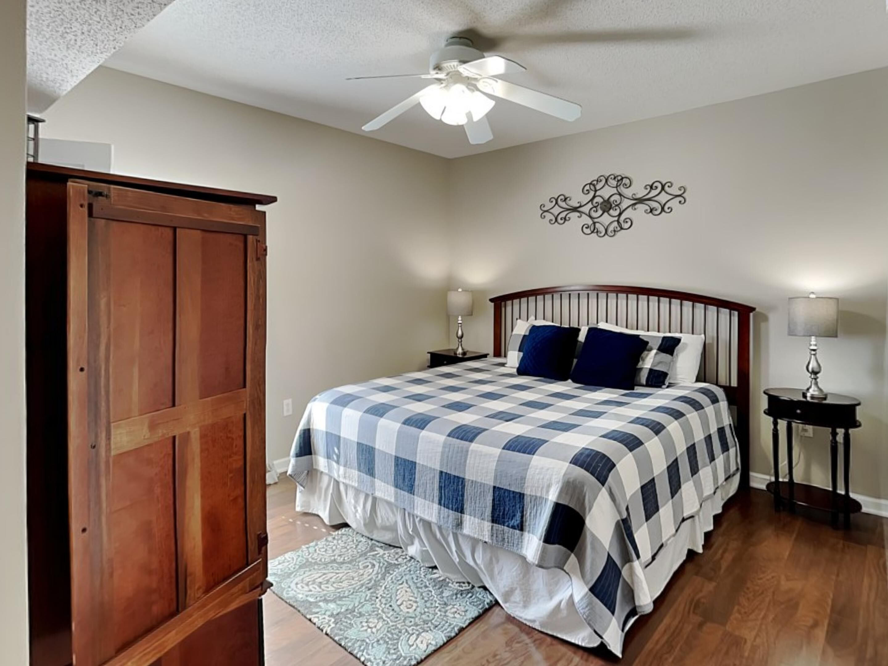 A bedroom with a checkered bedspread, a ceiling fan, a decorative wall piece, and a wooden wardrobe.