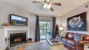 A living room with a fireplace, a television, a leather sofa, a coffee table, and a ceiling fan. - Bear Crossing by Jackson Mountain Rentals (Pigeon Forge)