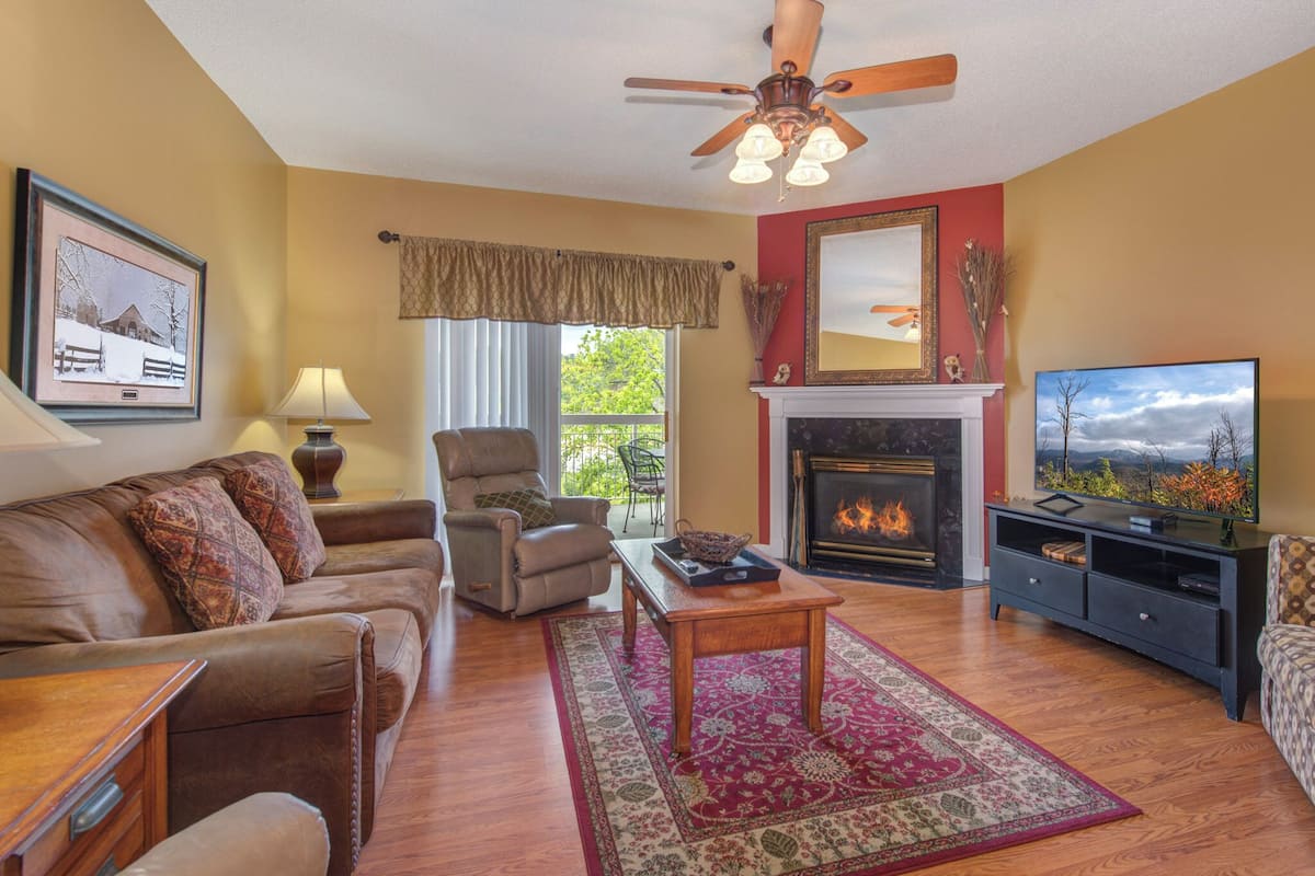 A living room with a fireplace, a ceiling fan, a television, and a large window with a view of trees.