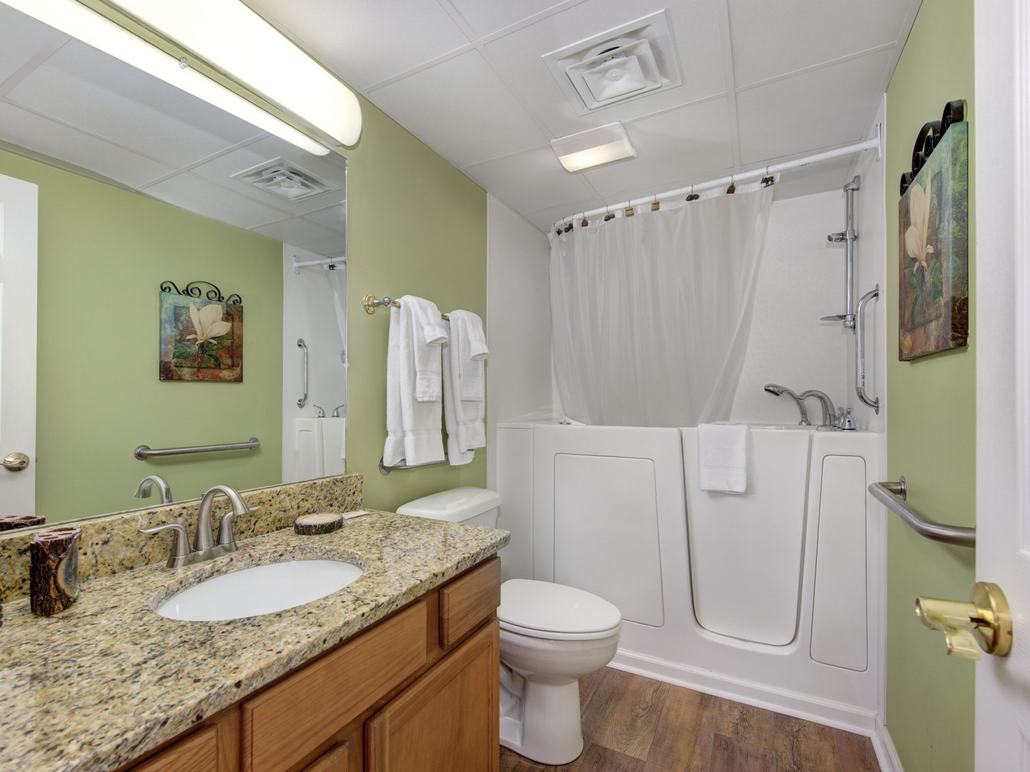 A bathroom with a granite countertop sink, a toilet, a bathtub with a shower curtain, and a handrail.