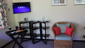 A room with a flat-screen TV displaying 'DSTV', a wicker chair with red cushions, a small table with a tray holding a glass of wine, and a framed picture on the wall.