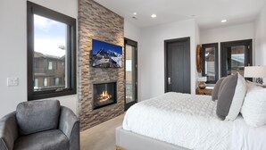 A modern bedroom with a fireplace, a large bed, a grey armchair, and a view of the outside.