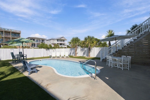 A swimming pool with a small hot tub, surrounded by white deck chairs and umbrellas, with a white fence and a staircase leading to a balcony.