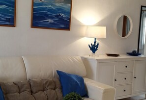 A living room with a beige sofa, a blue cushion, a white cabinet with drawers, a round mirror, and two framed paintings on the wall.