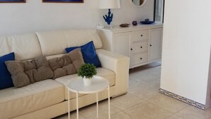 Una sala de estar con un sofá color beige, cojines azules, una pequeña mesa blanca con una planta, un mueble blanco con cajones, un espejo redondo y dos cuadros en la pared.