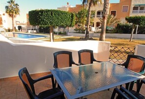 A terrace with a glass table and chairs, a swimming pool, and buildings in the background.