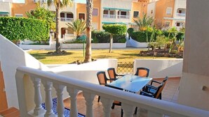 Un balcón con mesa y sillas, baranda blanca y vista a un patio con edificios y vegetación.