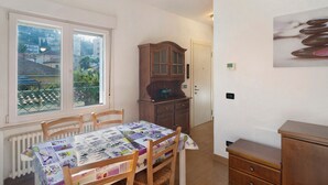 Uma sala de jantar com uma mesa coberta por toalha, cadeiras de madeira, um radiador, uma janela com vista para prédios e um armário.