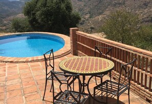 Ein Schwimmbecken mit einer gefliesten Kante, umgeben von einer Pflasterterrasse. Ein runder Tisch mit Stühlen und ein Metallzaun sind vorhanden.