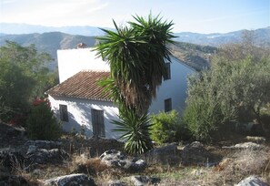 A white building with a tiled roof, surrounded by rocks and vegetation, with a mountainous backdrop. - Cottage "Sunny hillside casita in Guaro" with Mountain View, Pool, Terrace, A/C & Wi-Fi (Guaro)