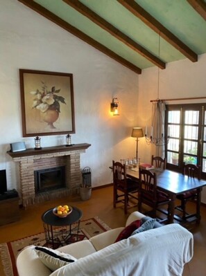 A cozy living room with a fireplace, dining table, and comfortable seating. - Cottage "Sunny hillside casita in Guaro" with Mountain View, Pool, Terrace, A/C & Wi-Fi (Guaro)
