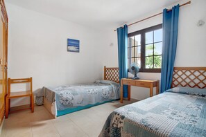 Une chambre avec deux lits, une chaise en bois, un bureau avec un ventilateur et une fenêtre avec des rideaux.