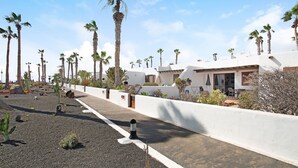 A pathway lined with palm trees, a white wall, and a building with a flat roof. - Apartment Puerto Chico R with Sea View, Pool, Terrace & Wi-Fi (Playa Blanca)