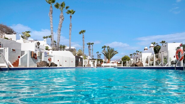 Una piscina con acqua cristallina, circondata da edifici bianchi e palme, sotto un cielo sereno.