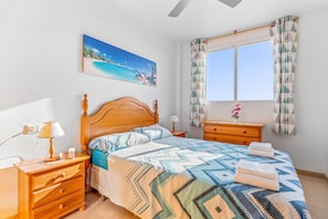 A bedroom with a wooden bed, a nightstand with a lamp, a dresser, a window with curtains, and a painting on the wall. - Apartment in Bolnuevo with Sea View, Shared Pool, A/C & Wi-Fi (Bolnuevo)