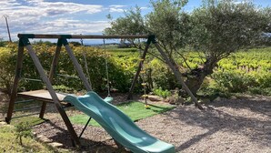 Eine Holzschaukel mit einer grünen Rutsche in einem Garten mit Olivenbäumen und einem Weinberg im Hintergrund.