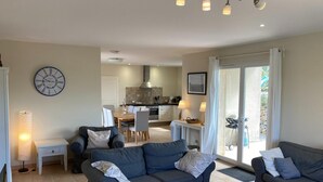 Ein modernes Wohnzimmer mit zwei dunklen Sofas, einem weißen Couchtisch und einem Essbereich mit Tisch und Stühlen. Im Hintergrund ist eine Küche zu sehen, an der Wand hängt eine große Uhr.