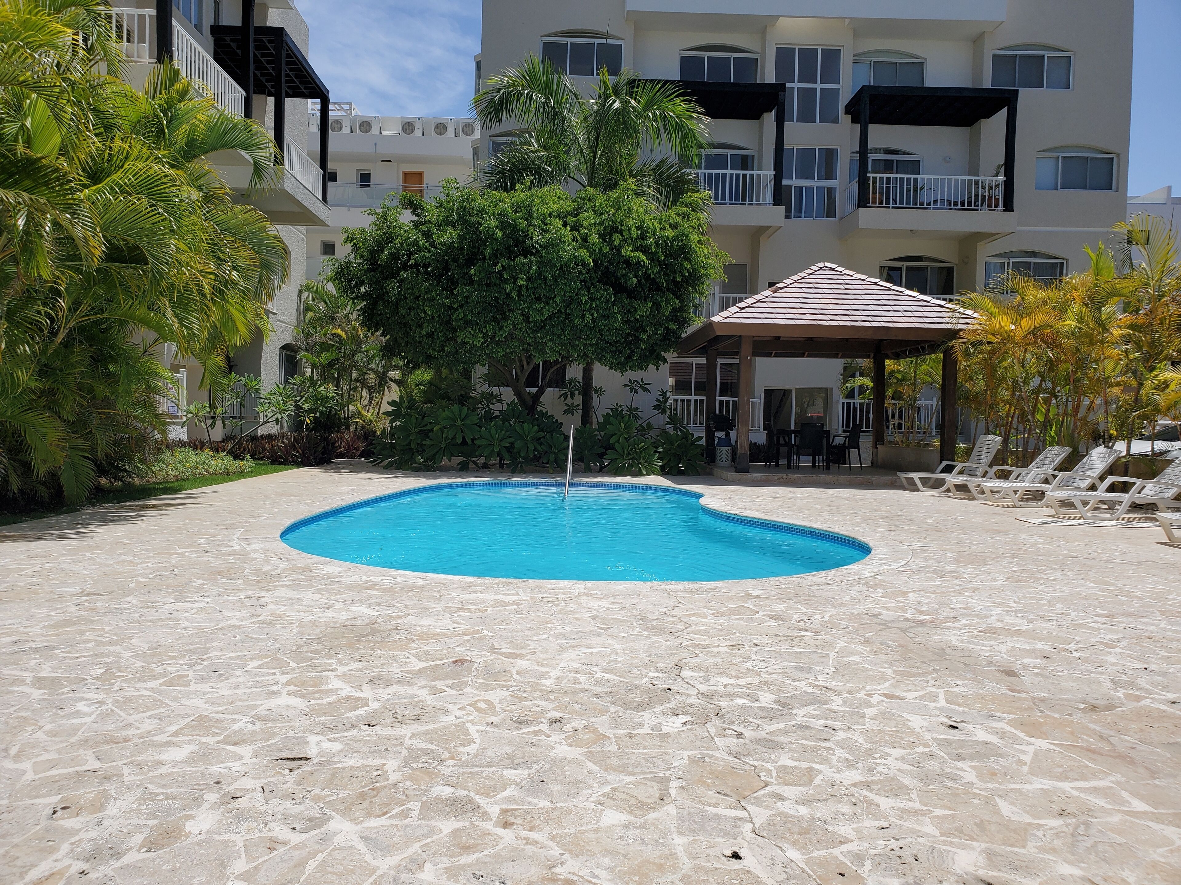 Una piscina rodeada de baldosas de piedra, con sillones de descanso y una zona de estar cubierta.