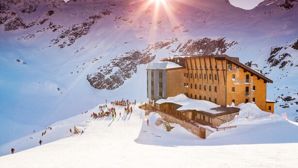 Eine Berghütte mit einem spiegelnden Teich davor, umgeben von schneebedeckten Bergen und einem klaren Himmel.