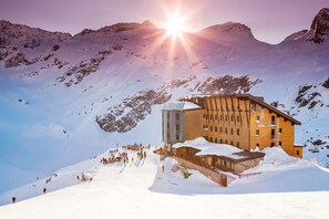 Un chalet de montagne avec un bassin aux reflets argentés devant, entouré de montagnes enneigées et surplombant un ciel dégagé.