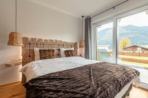 Un dormitorio con cabecera de piedra, una cama grande con manta marrón, una lámpara de noche y vistas a montañas a través de un ventanal amplio.