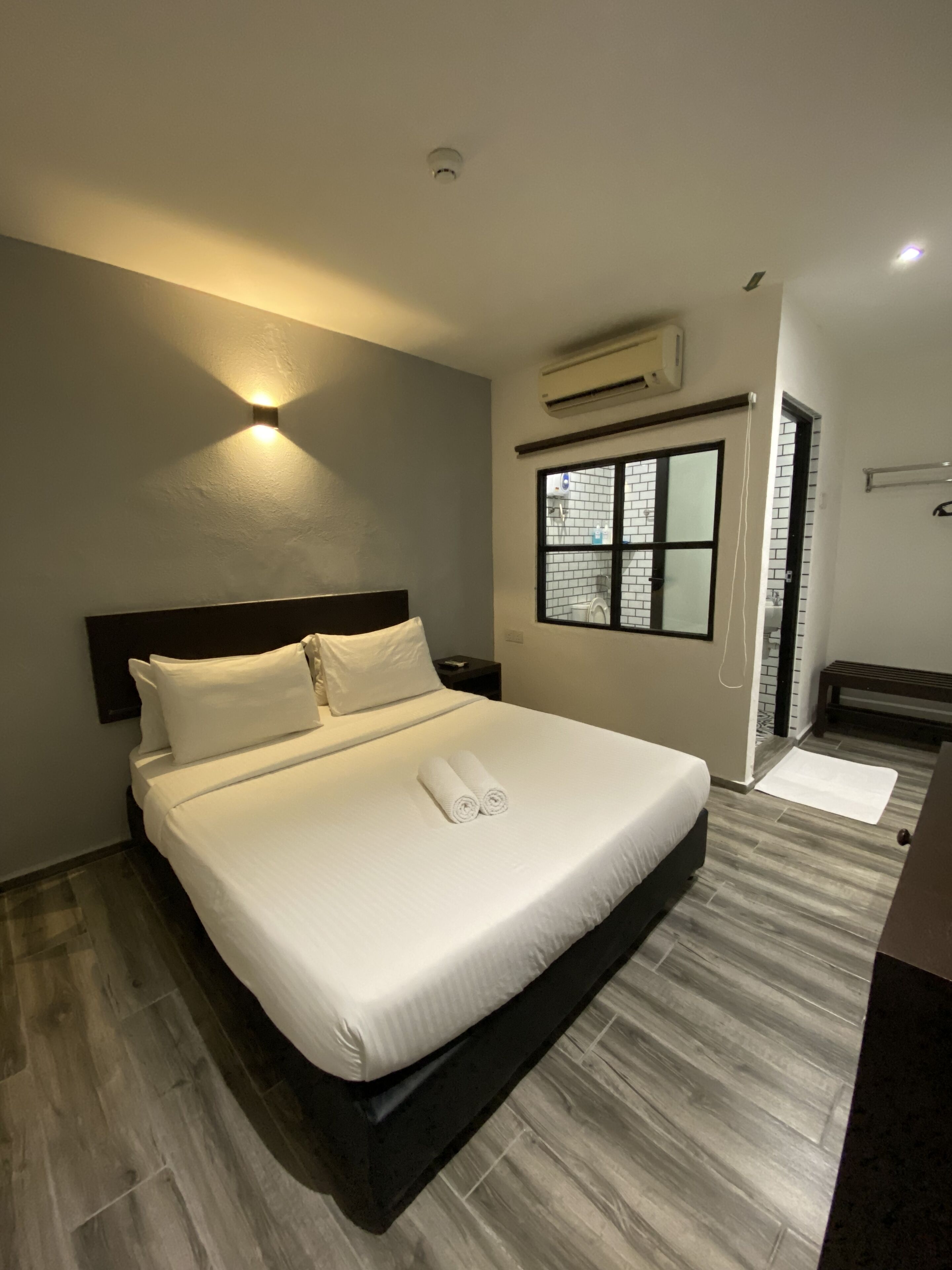 A modern hotel room with a large bed, a nightstand, a window with a view of the outside, and a built-in air conditioning unit.
