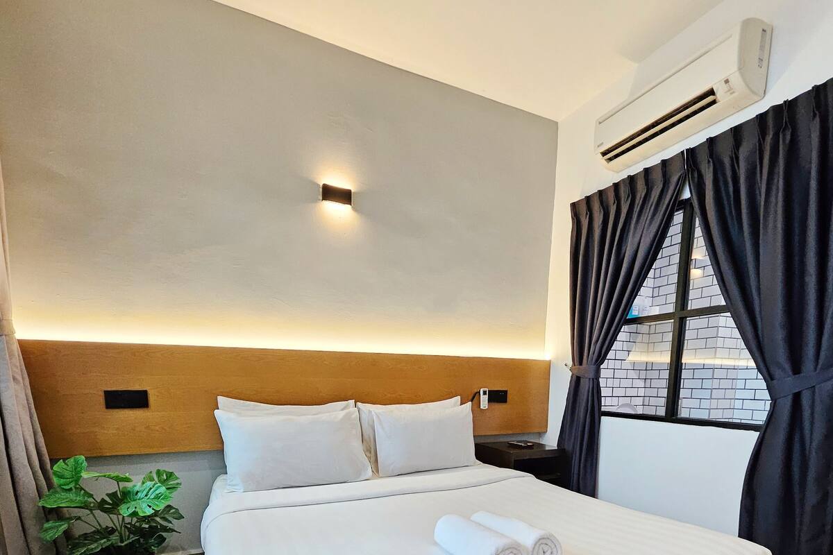Ein Hotelzimmer mit einem großen Bett, einem Blumentopf, einer Wand-Klimaanlage und einem mit Vorhängen versehenen Fenster.