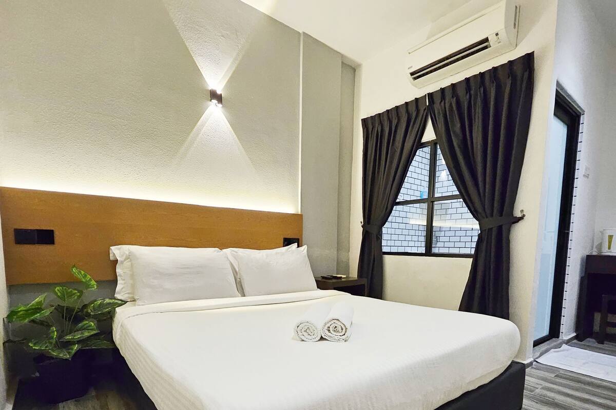Ein Hotelzimmer mit einem großen Bett, einer Wand-Klimaanlage, einem Topf mit Pflanze und einem fensterbepflanzten Vorhang.