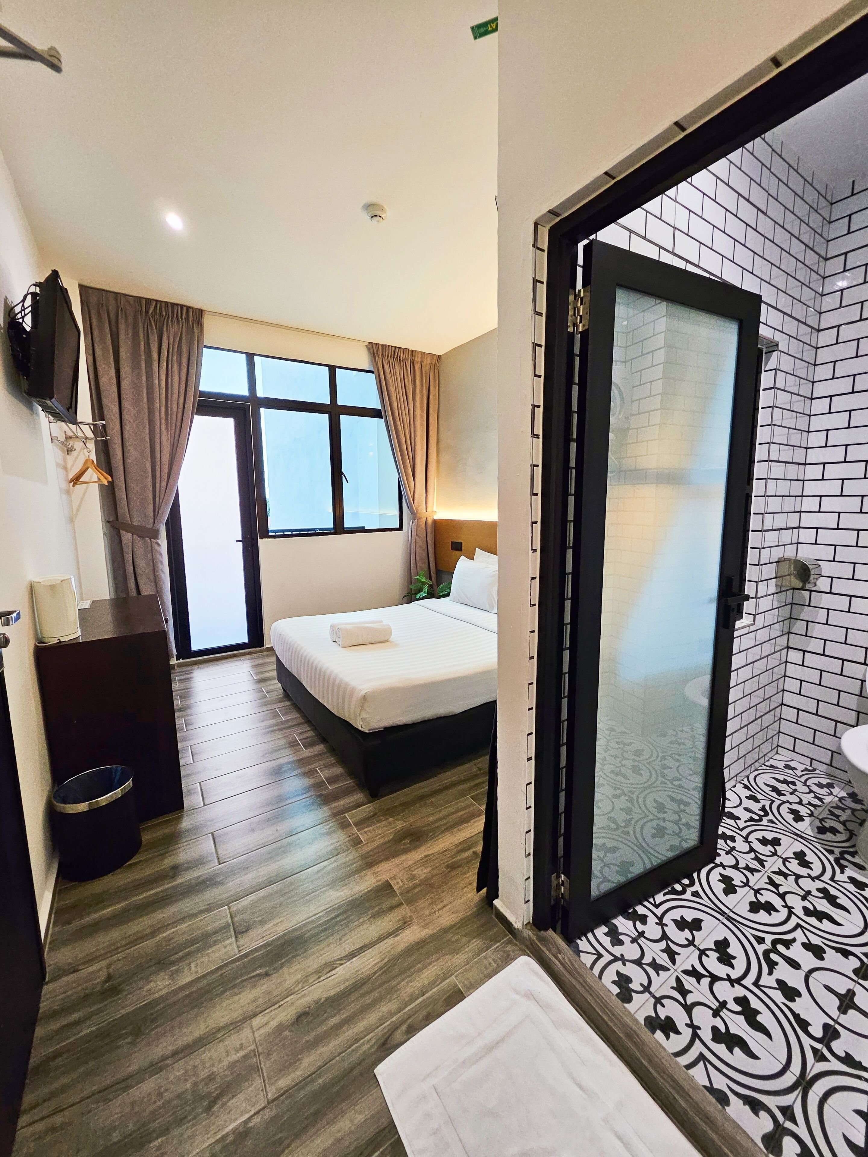 A hotel room with a bed, a desk, a TV, a window with curtains, and a bathroom with a tiled floor and a mirror.