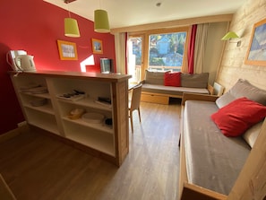 A compact living space with a kitchenette, a sofa with red cushions, and a window offering a view of a residential area.
