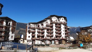 Un hôtel à plusieurs étages doté de balcons, un panneau indiquant la présence d’une station de ski, et des montagnes en arrière-plan.