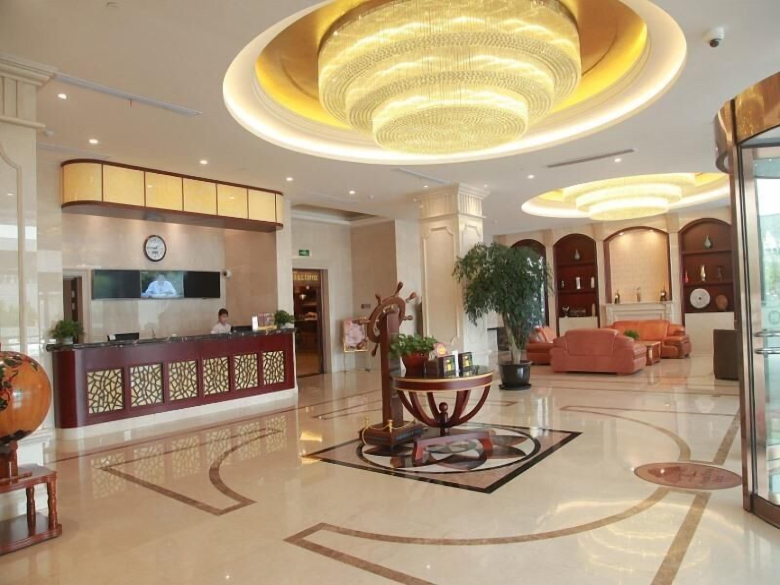A hotel lobby with a reception desk, a large chandelier, a seating area with sofas, and a decorative table with plants.