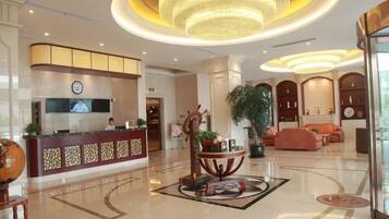 A hotel lobby with a reception desk, a large chandelier, a seating area with sofas, and a decorative table with plants.