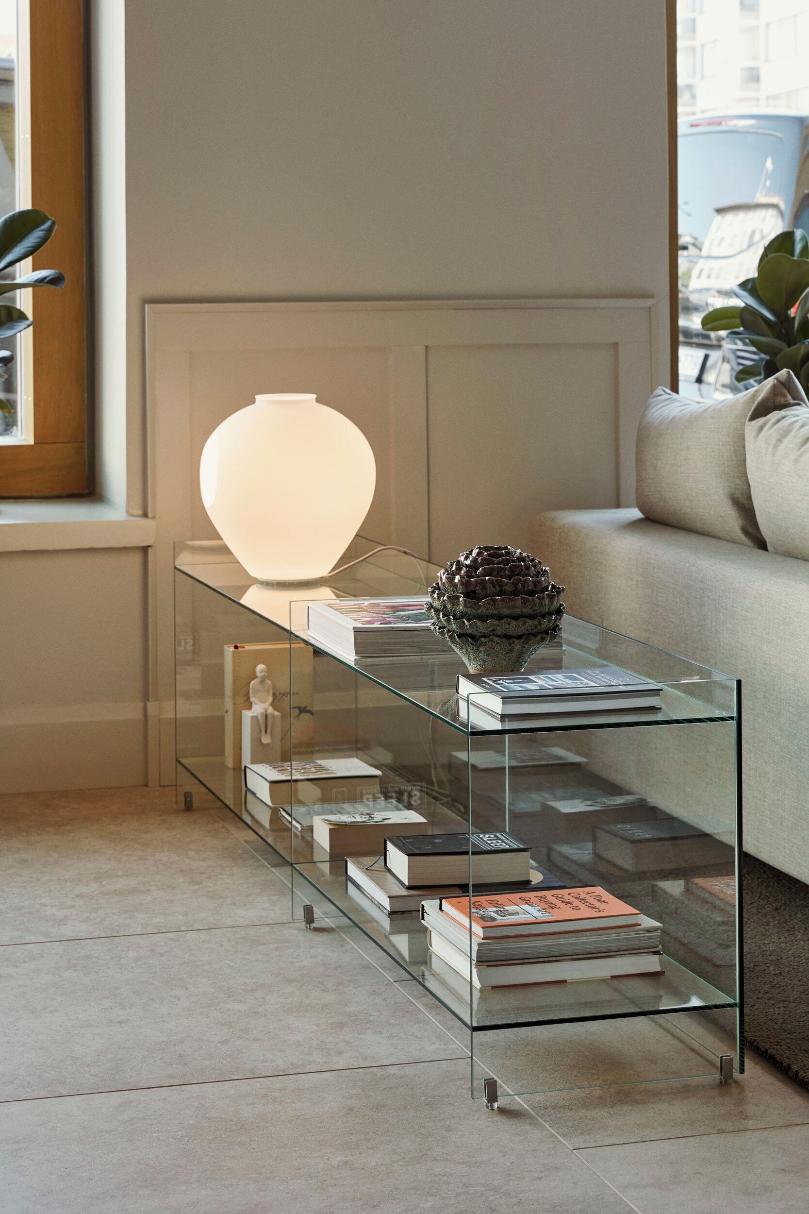 A modern living room with a glass coffee table displaying books and a decorative object, a sofa, and a window with a view of the city.