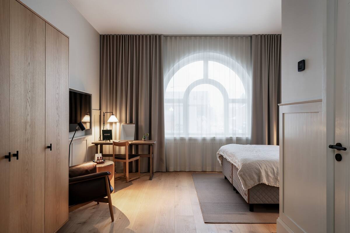 A hotel room with a bed, a desk with a lamp, a chair, a wardrobe, and a large window with curtains.