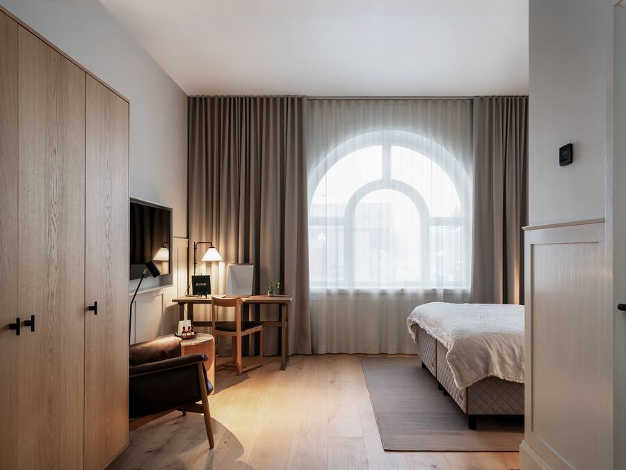 Ett hotellrum med en säng, ett skrivbord med en lampa, en stol, en garderob och ett stort fönster med gardiner.