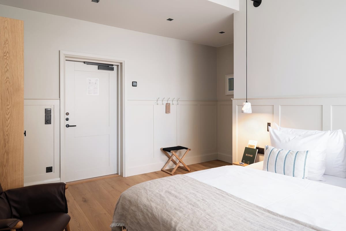 A hotel room with a bed, a wooden chair, a small stool, a nightstand, and a door with a sign.