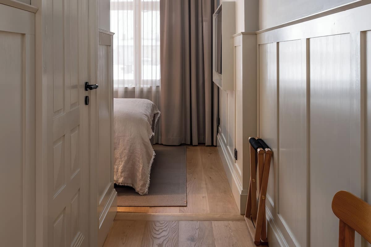 A narrow hallway with a wooden floor leading to a bedroom with a bed and curtains.