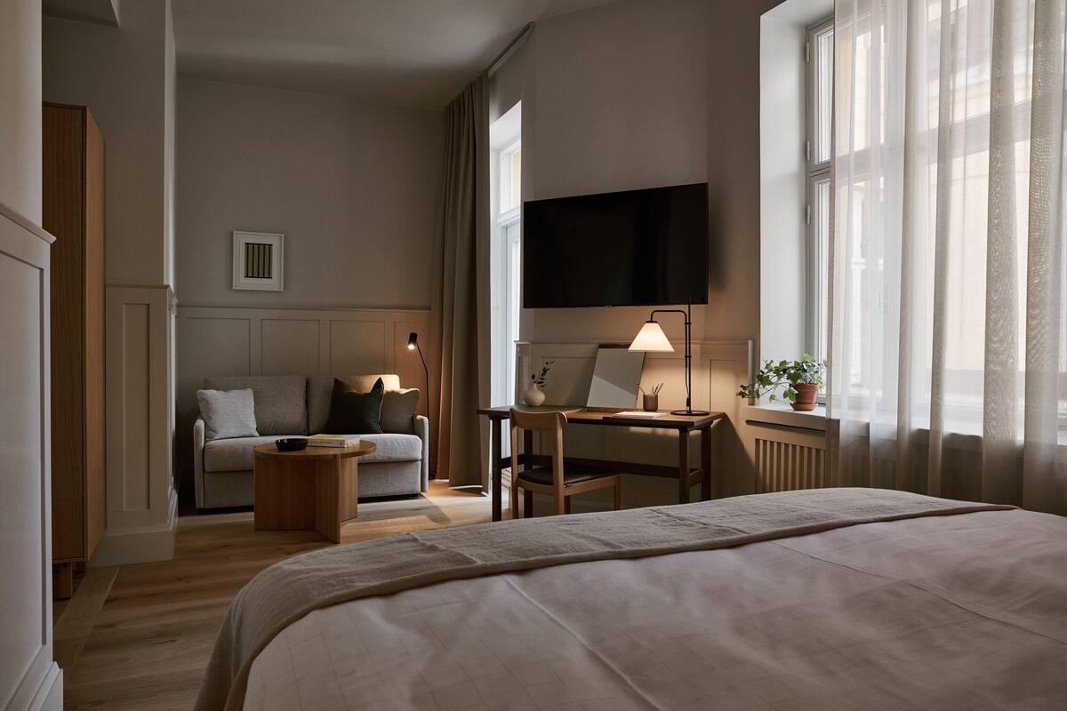 A hotel room with a bed, a desk with a lamp, a chair, a sofa, and a TV mounted on the wall.