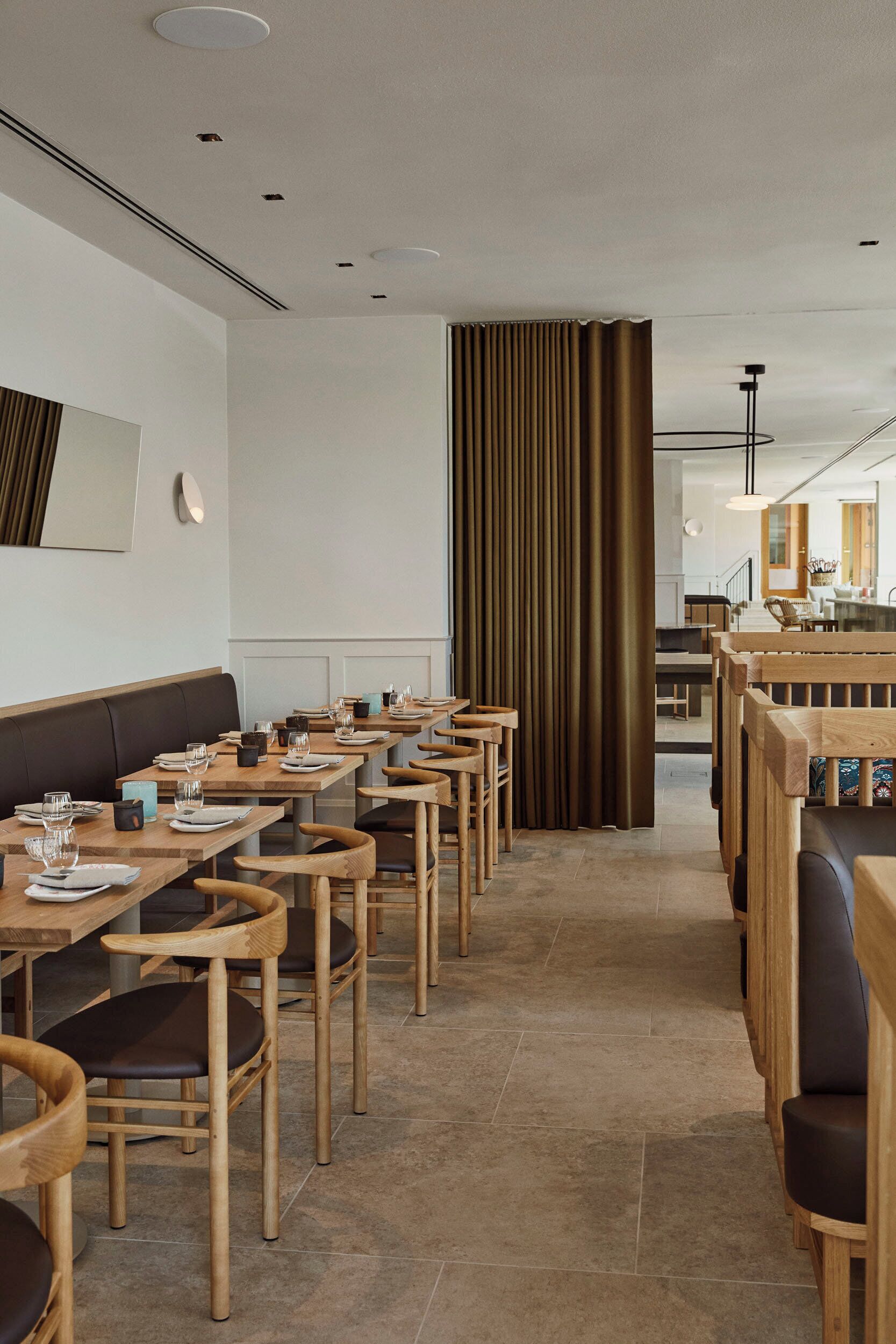 A modern restaurant interior with wooden tables and chairs, a central aisle, and a view into the kitchen area.