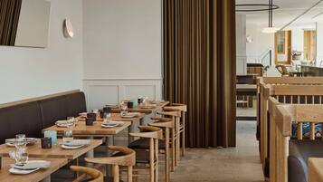A modern restaurant interior with wooden tables and chairs, a central aisle, and a view into the kitchen area.