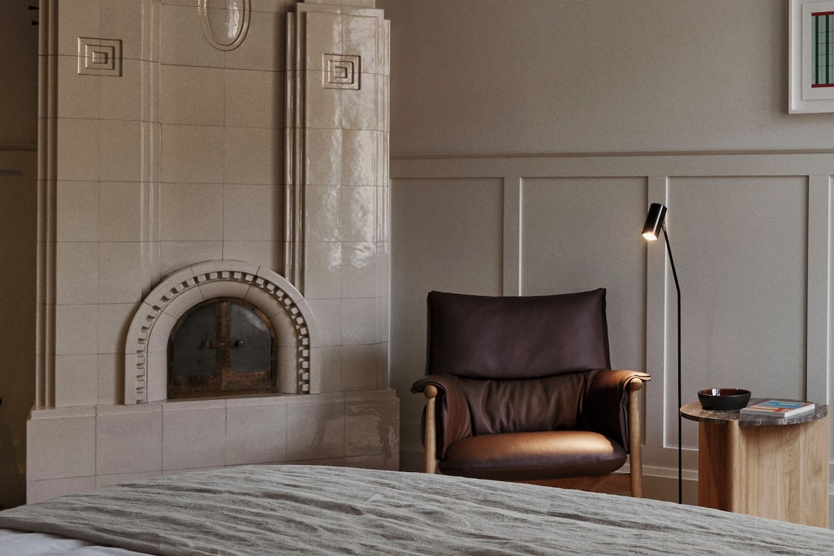 A bedroom with a large bed, a fireplace, a leather chair, and a bedside table with a lamp.